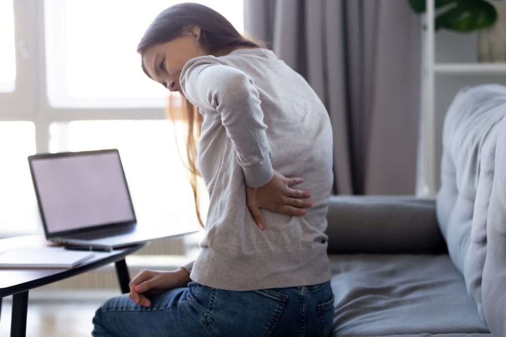 Young woman experiencing back pain after sedentary computer work sitting in bad posture on couch. 